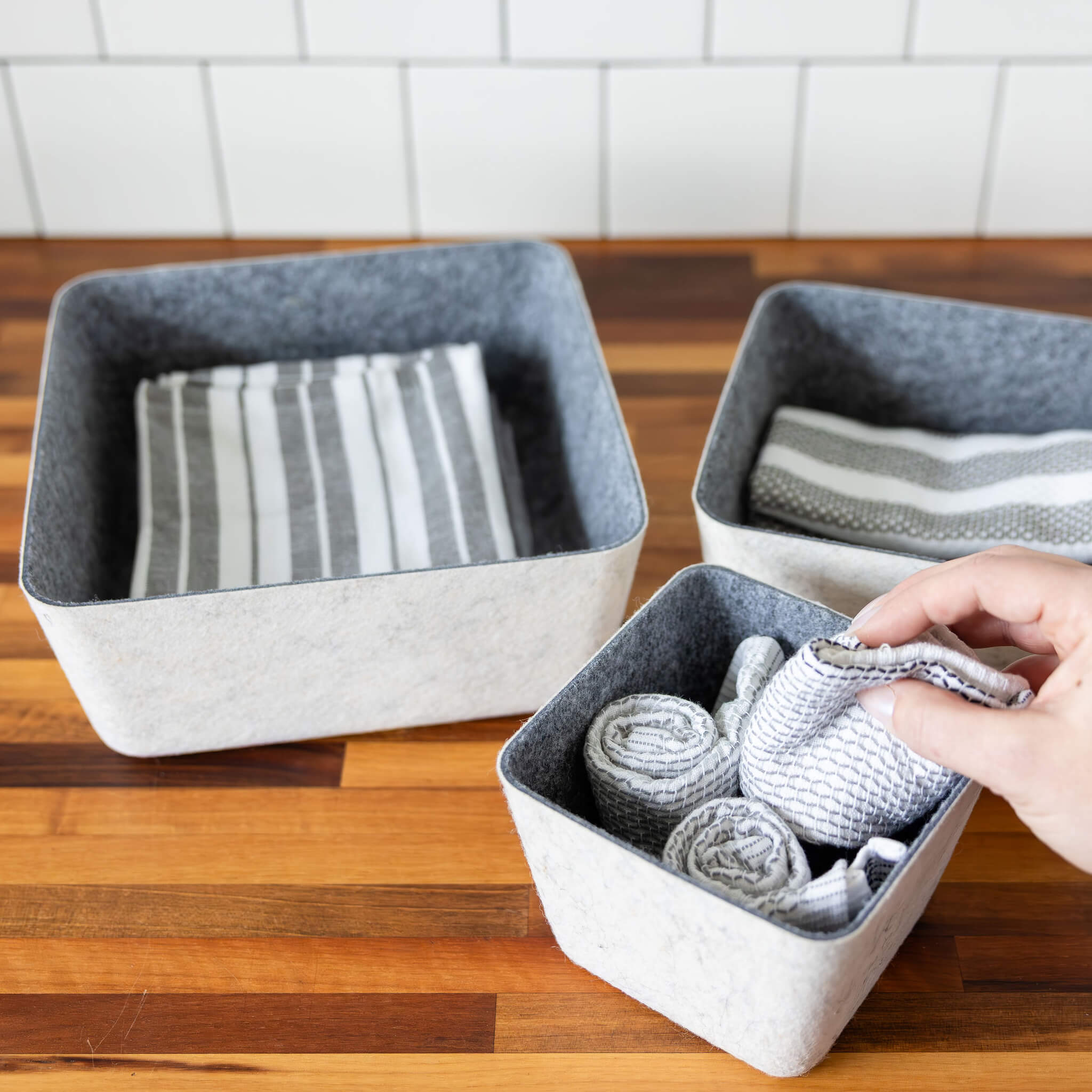 Three square recycled felt organizing sorting storage bins with charcoal interiors and cream exteriors on a wooden surface with a tiled wall in the background.