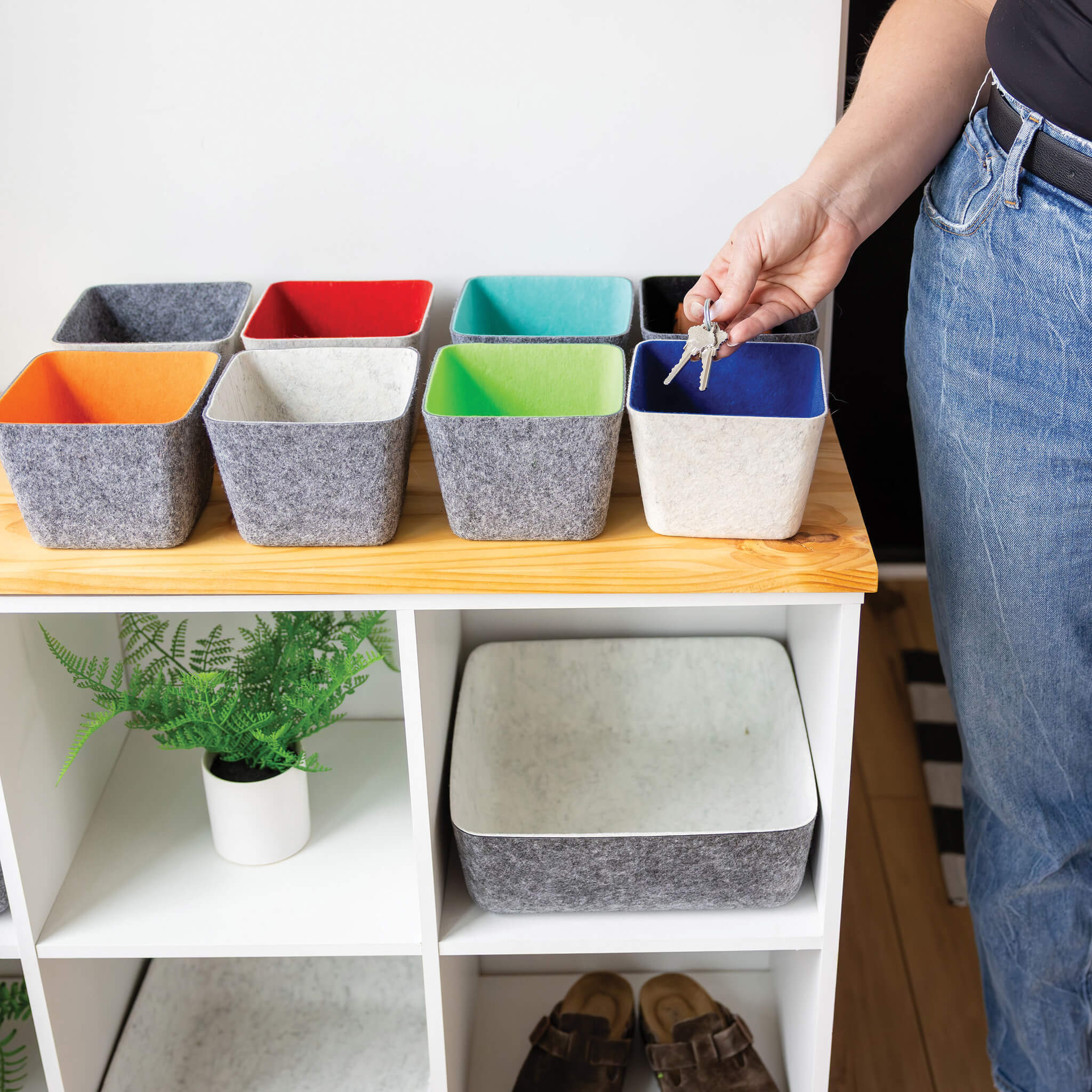 Colorful recycled felt organizing sorting storage bins on a wooden shelf with a person putting keys into one, on a white cabinet with a plant and shoes.
