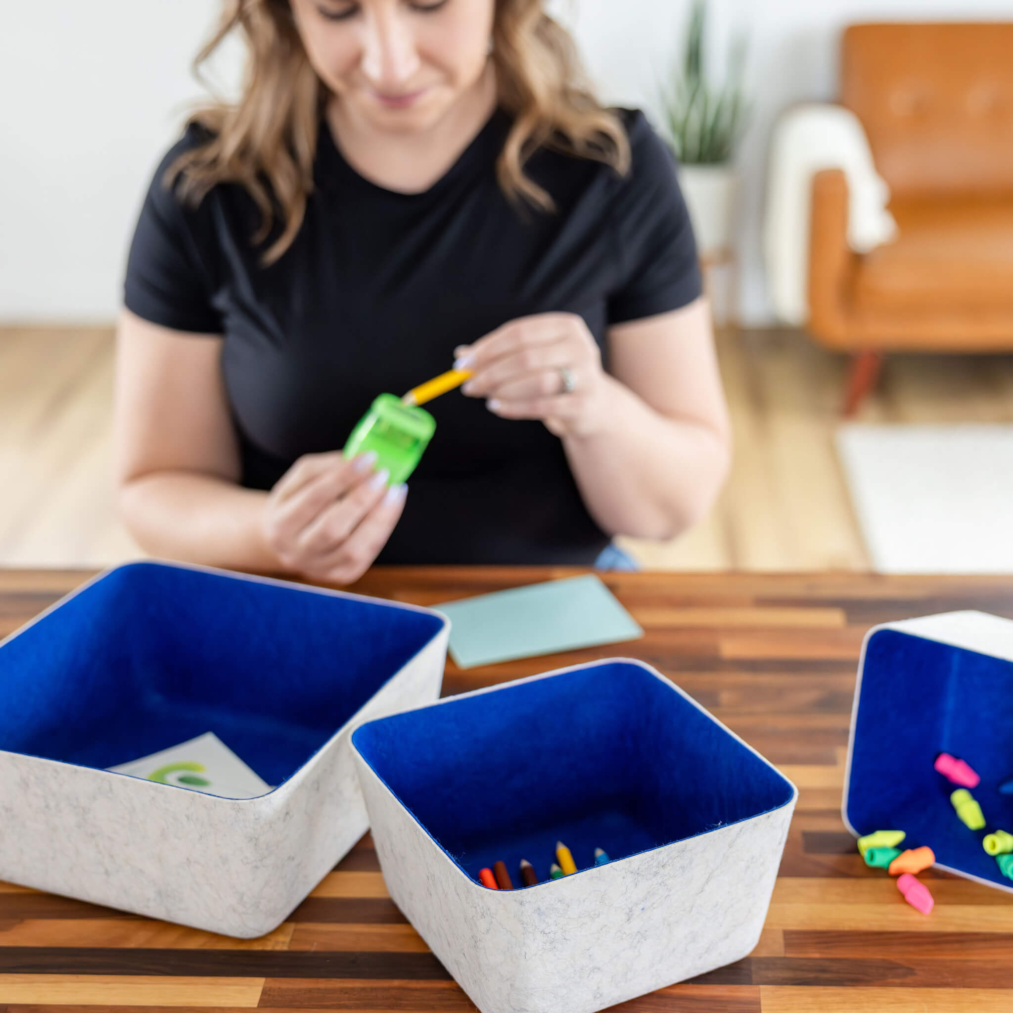 Woman organizing art supplies into recycled felt organizing sorting storage bins with navy interior and cream exterior on a wooden table.