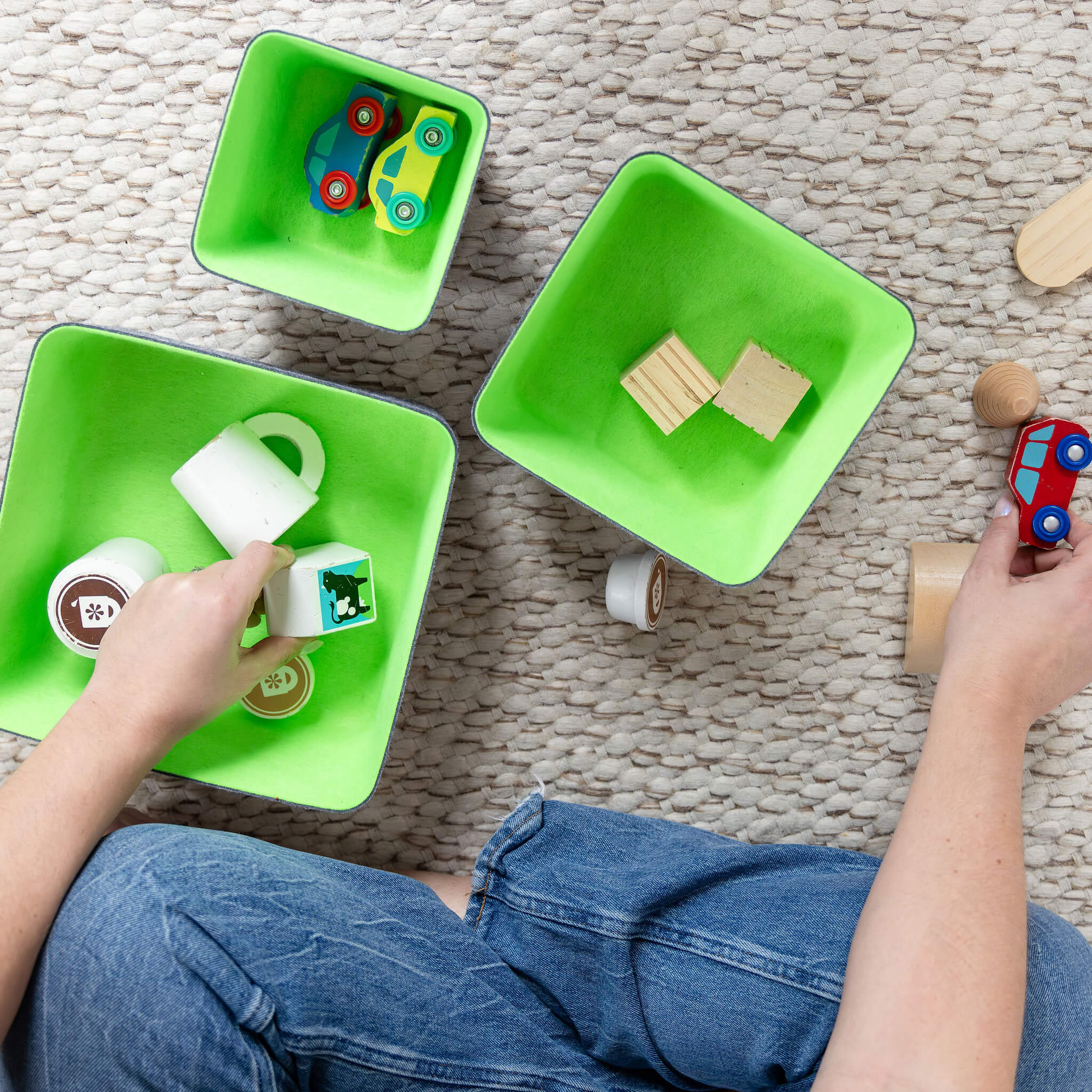 Person organizing  toys into recycled felt organizing sorting storage bins with green interior and grey exterior on a textured floor.